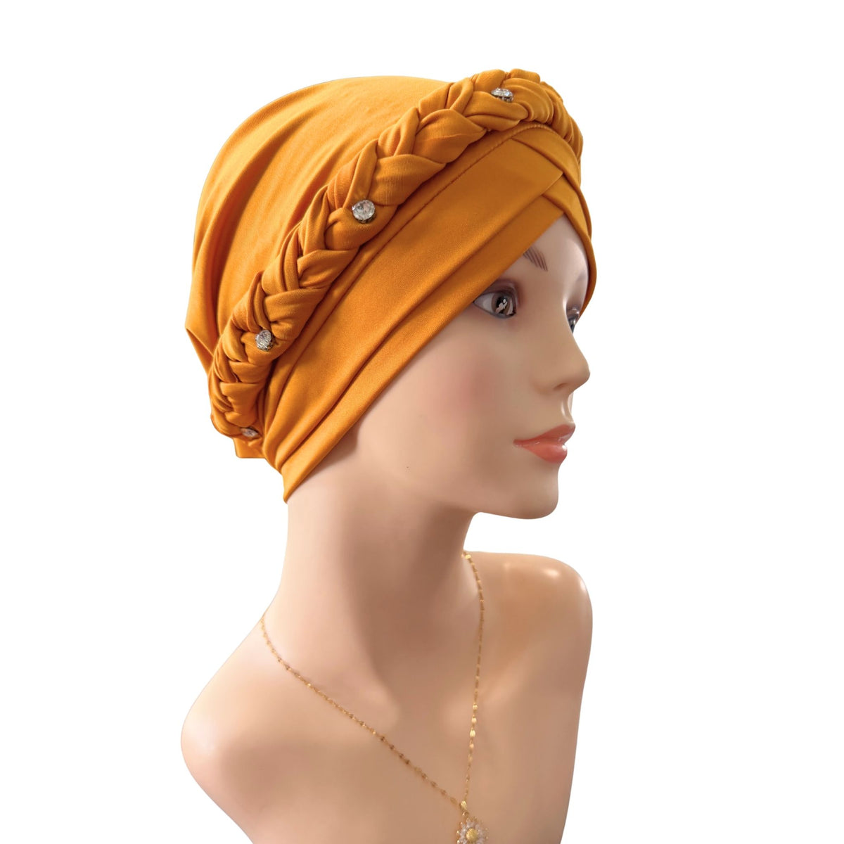 A mustard yellow fabric headwrap or turban hat with braided detailing and small rhinestone embellishments, displayed on a mannequin head against a plain white background.
